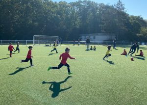 Kinder in roten und gelben Trikots spielen auf dem Kunstrasen auf Minitore, vorne läuft ein Junge im roten Trikot zum Ball